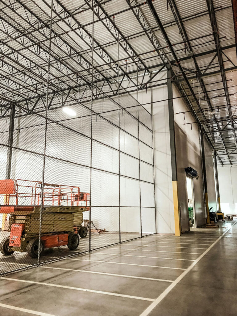 A spacious warehouse with high ceilings, metal beams, and a chain-link fence. Lift equipment is parked inside, and the area is well-lit with overhead lights and clearly marked parking spaces.