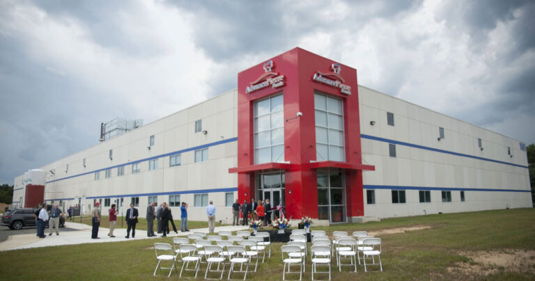 A group of people stand and talk outside a large white building with a bold red entrance, surrounded by empty white chairs arranged in rows on a grassy lawn, under a cloudy sky.