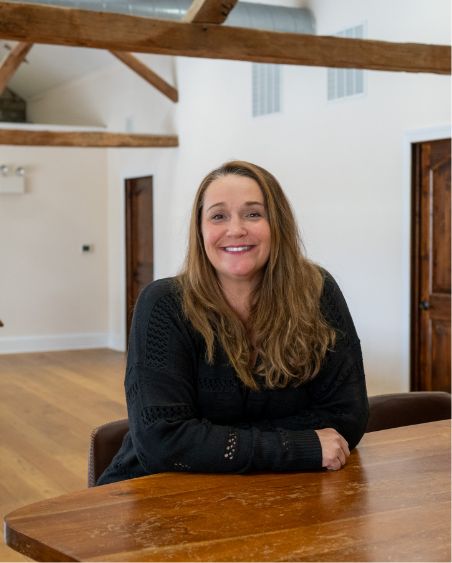 A woman with long brown hair, wearing a black sweater, sits at a wooden table in a bright room with wooden beams, white walls, and wooden doors, smiling at the camera.