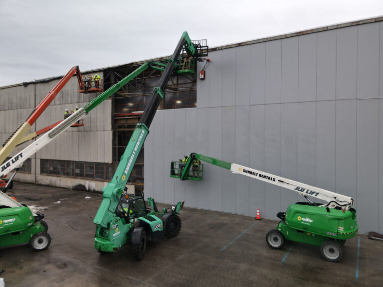 Construction workers using green boom lifts and cherry pickers to work on the exterior walls and windows of an industrial building. The ground is wet and several workers are visible wearing safety gear.