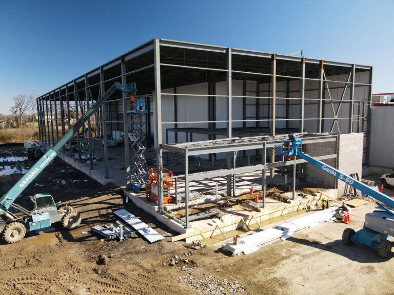 A large building under construction, with a steel framework partially assembled. Construction equipment and cranes are on site, and workers can be seen working on the structure under a clear blue sky.
