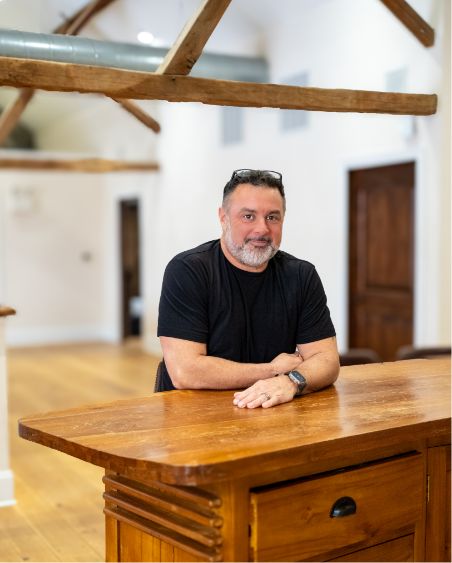 A man with short hair and a beard, wearing a black t-shirt and glasses on his head, sits at a wooden counter in a bright room with wooden beams and light-colored floors.