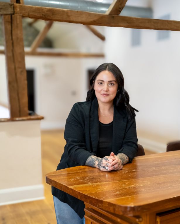 A woman with dark hair, wearing a black jacket and jeans, stands leaning on a wooden table in a bright room with exposed wooden beams and white walls. She has visible tattoos on her hands and is smiling gently at the camera.
