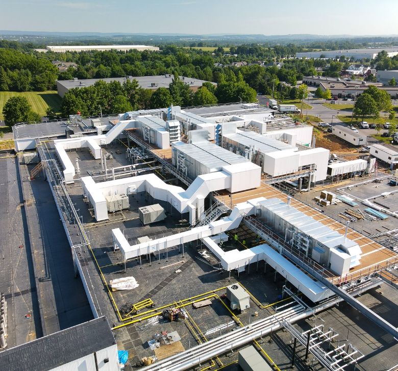 Aerial view of a large industrial facility with extensive white ventilation ducts, equipment, and interconnected buildings, surrounded by green trees, fields, and additional structures in the background under a clear sky.