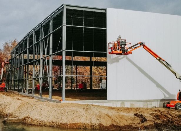 A construction worker on a cherry picker installs white panels on the exterior frame of a large metal building, surrounded by construction equipment and autumn trees.