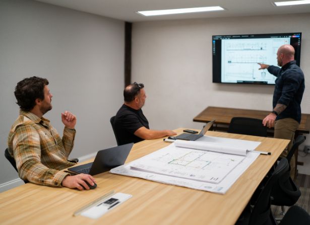 Three men in a meeting room, two seated at a table with laptops and blueprints, while one stands and points at architectural plans displayed on a large screen.