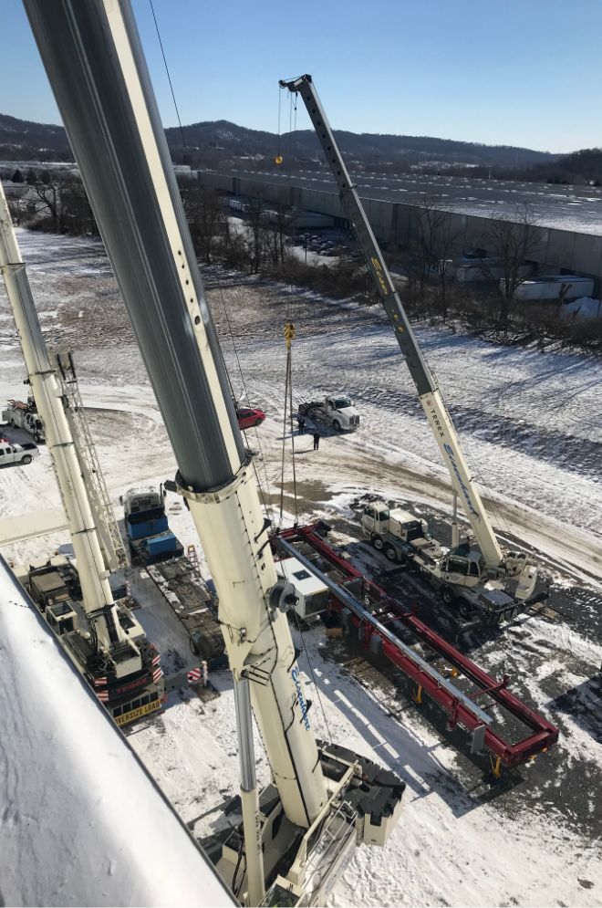 Three large mobile cranes operate on a snowy construction site, lifting and positioning a long red steel beam near a flatbed truck. Industrial buildings and snowy landscape are visible in the background under a clear sky.