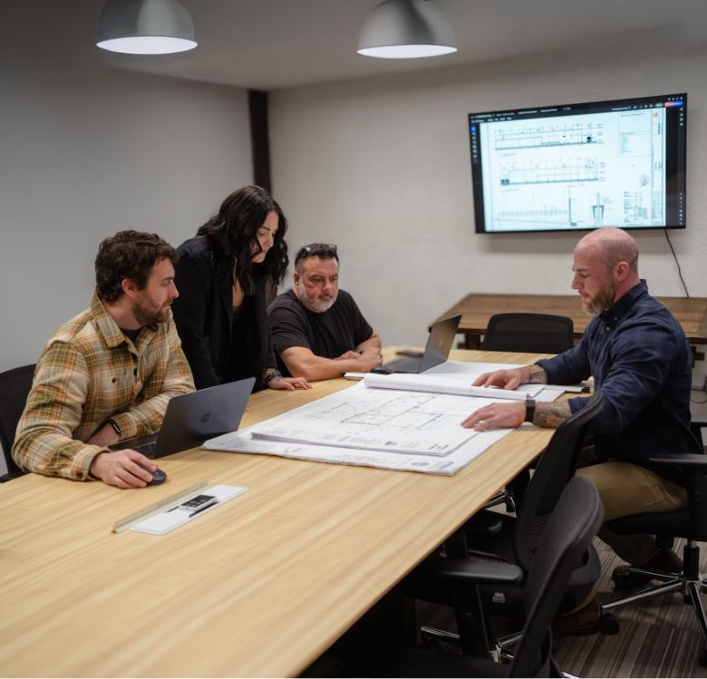 Four people sit around a conference table reviewing architectural blueprints. Two men use laptops, while another man and a woman look at prints. A monitor on the wall displays a technical drawing.