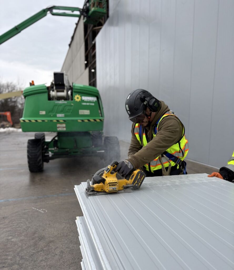 A construction worker wearing safety gear and a reflective vest uses a power saw to cut large metal panels outdoors near a gray industrial building and a green lift.