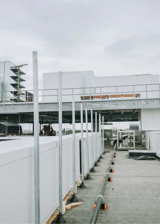 A rooftop with industrial HVAC equipment, white units, metal railings, vertical poles, and visible conduit lines under an overcast sky.