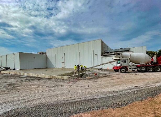 A group of construction workers stand near a cement truck pouring concrete outside a large white industrial building under a partly cloudy sky.