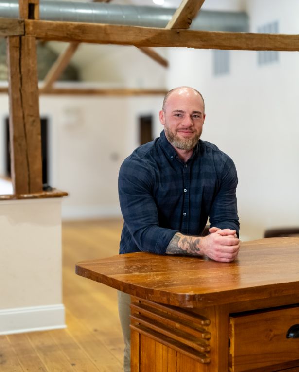 A man with a shaved head and beard, wearing a dark plaid shirt, stands behind a wooden desk in a bright room with exposed beams and wooden floors, smiling at the camera with arms resting on the desk.