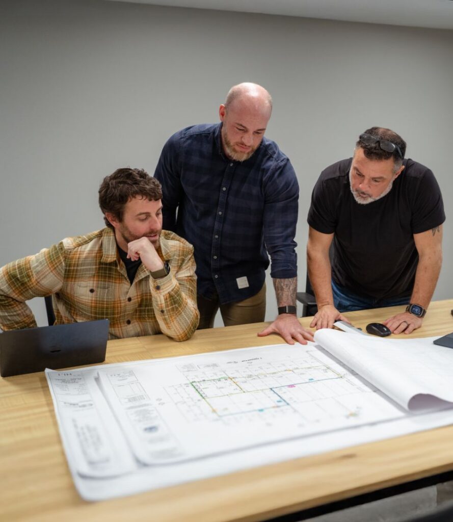 Three men stand and sit around a table, looking over large architectural blueprints. One man points at the plans while another sits with a laptop, and a third observes, all focused on the documents.