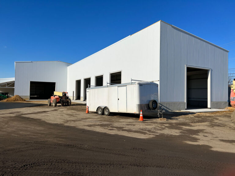 A large white industrial warehouse with several open garage doors, a white trailer, an orange forklift, and traffic cones in the foreground on a sunny day.