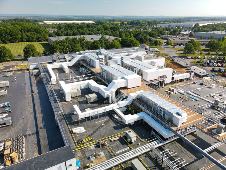 Aerial view of a large industrial facility with interconnected white structures, pipes, and ventilation systems, surrounded by trees, parking lots, and nearby buildings under a clear sky.