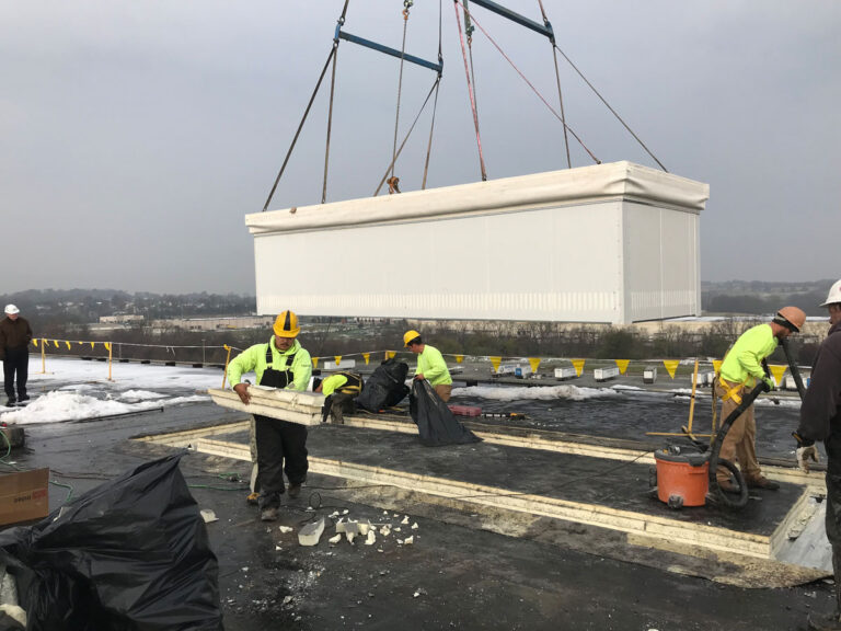 Workers in safety gear remove debris from a rooftop while a large white prefabricated structure is being lifted by a crane in the background. Yellow caution tape surrounds the work area.