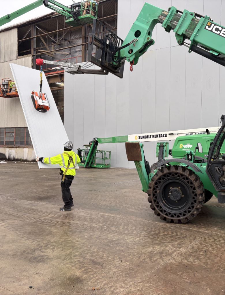 A construction worker guides a large metal panel being lifted by a green forklift with Sunbelt Rentals branding outside an industrial building. Another machine stabilizes the top of the panel.