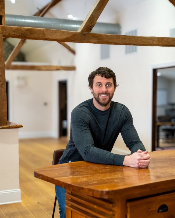 A man with curly brown hair and a beard, wearing a dark long-sleeve shirt, sits smiling at a wooden desk in a bright room with exposed wooden beams and hardwood floors.