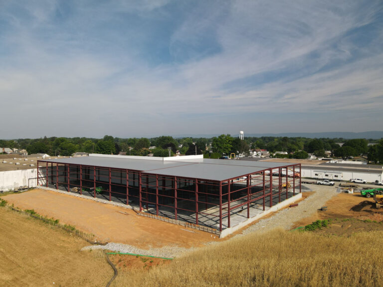 Aerial view of a large commercial building under construction, showing a steel frame structure with a partially completed roof, surrounded by dirt, grass, and nearby buildings under a blue sky.