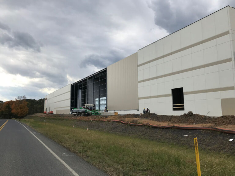A large industrial building under construction beside a road, with construction equipment and workers visible near the open section of the structure under a cloudy sky.