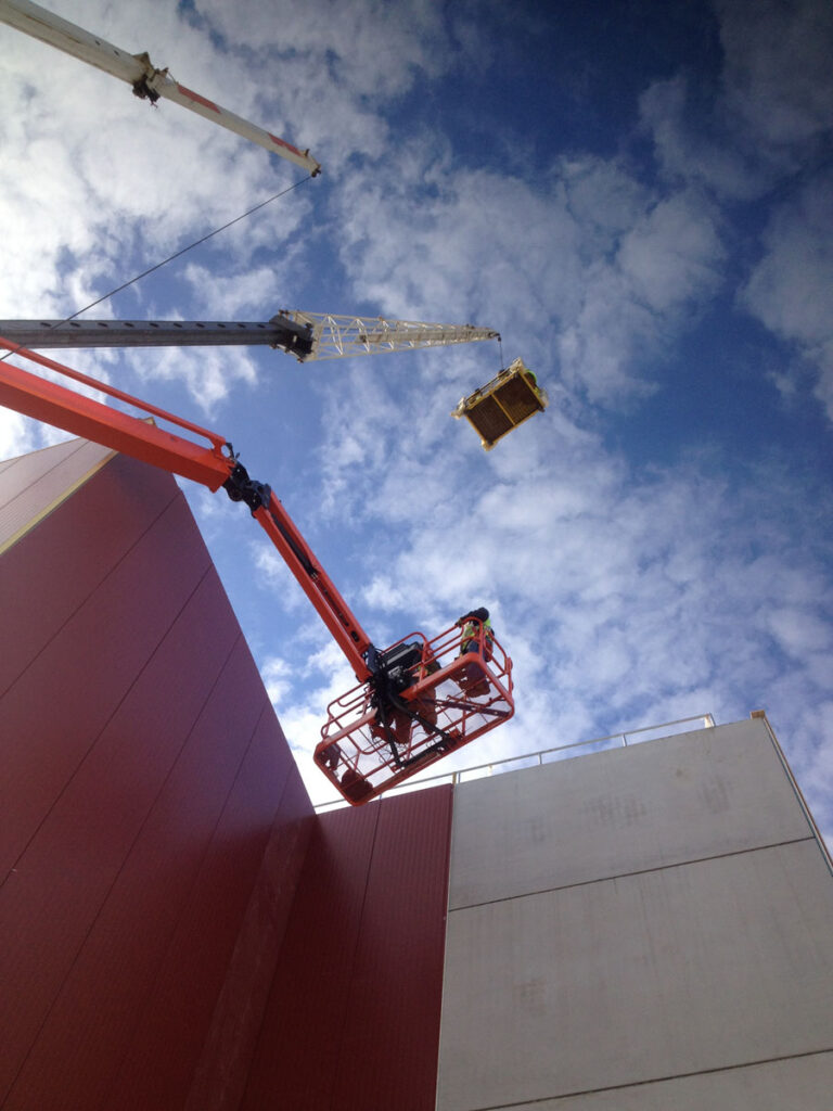 A worker stands on an orange boom lift next to a tall building while a crane hoists a platform overhead against a blue sky with scattered clouds.
