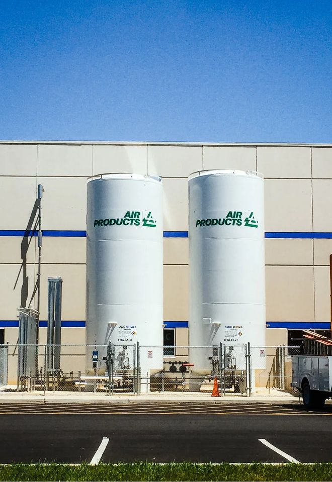 Two large white industrial tanks labeled Air Products stand side by side outside a beige building with blue trim, surrounded by a chain-link fence and equipment, under a clear blue sky.