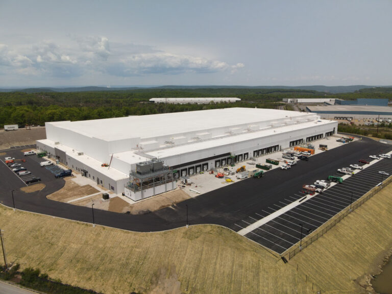A large white industrial warehouse with loading docks and several trucks and cars parked outside, surrounded by greenery and a mostly empty parking lot, viewed from above.
