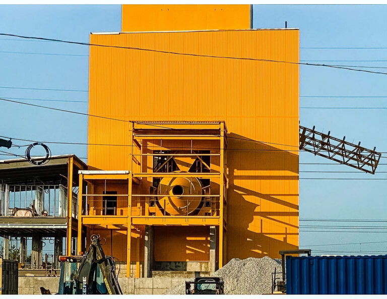 A bright yellow industrial building with a large circular fan on its facade, construction materials, and equipment in front. Power lines cross the image and a conveyor extends from the right side of the building.