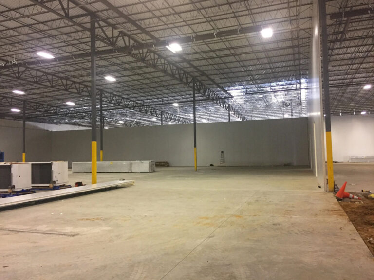 Large, empty warehouse with concrete floor, high ceiling, exposed beams, and yellow safety bollards. Construction materials and equipment are scattered around, with bright overhead lights illuminating the space.