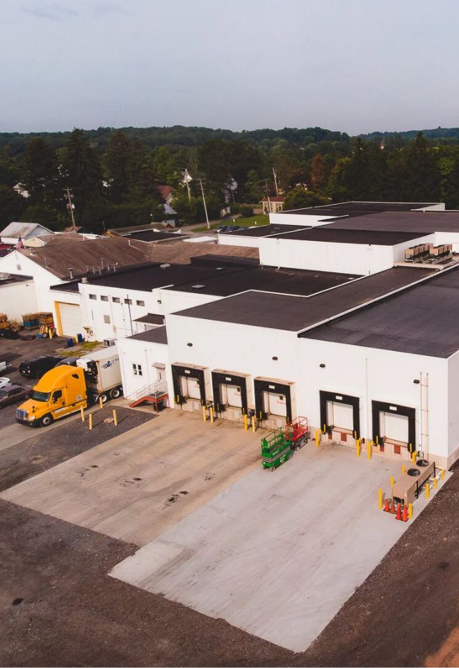 Aerial view of a white industrial warehouse with four loading docks, a yellow semi-truck parked nearby, and trees and houses in the background.