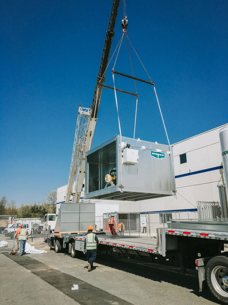 A crane lifts a large metal container onto a flatbed truck outside an industrial building, while workers in safety vests and hard hats guide the operation under a clear blue sky.