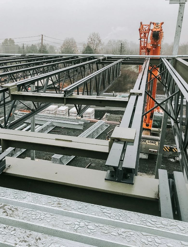 Steel beams and framework at a construction site, with an orange crane in the background and stacks of materials on the ground, all under a cloudy sky.