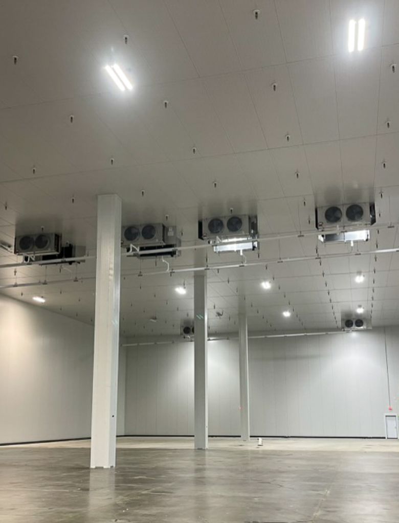 A large, empty industrial warehouse with high ceilings, evenly spaced support columns, bright overhead lighting, and multiple ceiling-mounted ventilation or refrigeration units. The floor appears clean and unfinished.