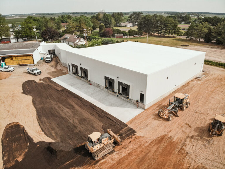 Aerial view of a large, white warehouse building with multiple loading docks. Construction vehicles and workers are paving the surrounding dirt area. Nearby houses and trees are visible in the background.