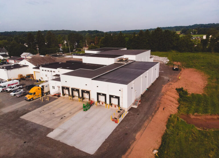 Aerial view of a large white industrial warehouse with multiple loading docks, trucks parked nearby, and surrounding greenery and trees in the background.