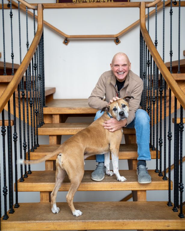 A smiling man sits on wooden stairs inside, petting a tan and white dog that stands in front of him. The man wears a brown jacket and blue jeans, and the background features black iron stair railings.